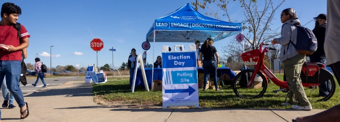 Students walking past white and blue signs for Election Day Polling sites and a blue Student Engagement tent on a sunny fall day.