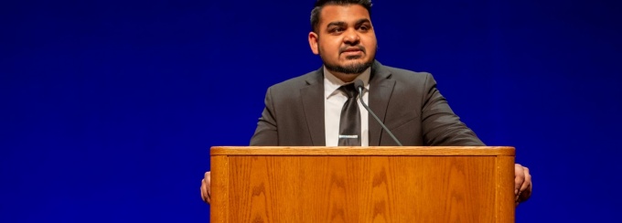 Former SA President Nicholas Singh wears a dark grey suit while standing at a wooden podium with the interlocking UB logo on the front. The background is uplit in a bright UB blue during a speaking engagement.