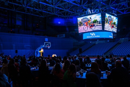 Climate Action Plan Event on the basket ball court inside alumni areana with illuminated jumbotron and blue lights with people in seats and a speaker on the stage.