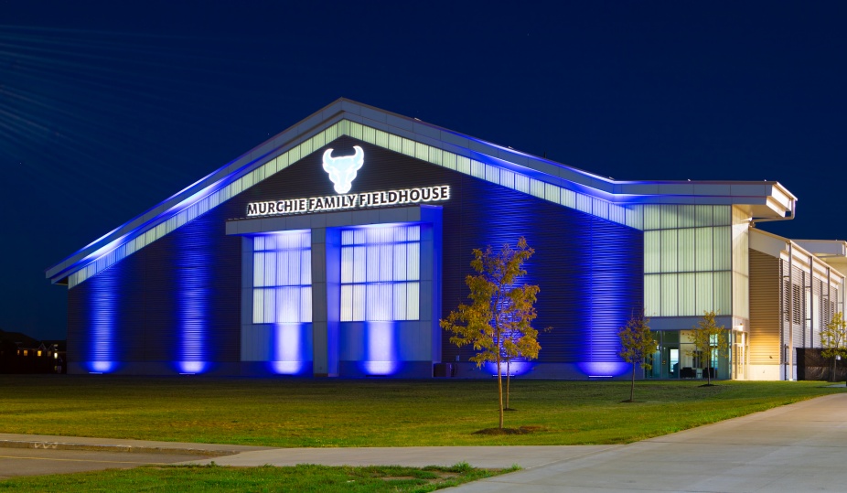 Exterior of the Murchie Family Fieldhouse at night. The front of the building is lit up in blue. 