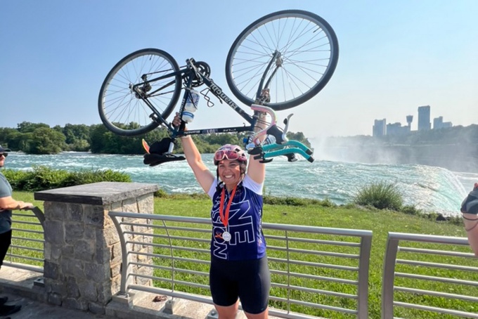 Emily Genovesi holds her bike over her head, Niagara Falls in the background. 