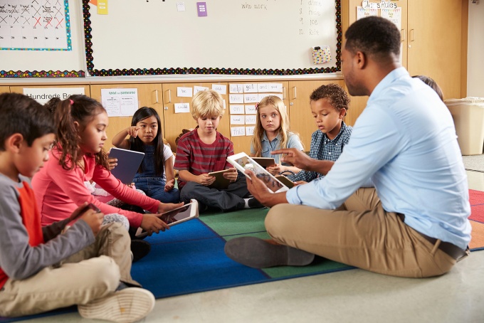 Male teacher reading to young kids sitting around him on the floor. 