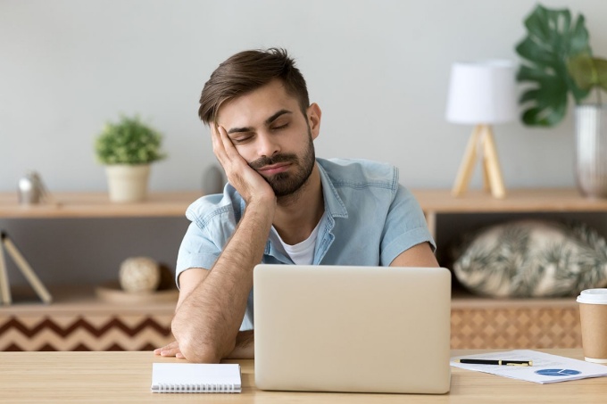 Young man sitting at his laptop, head leaning on one hand, eyes closed. 